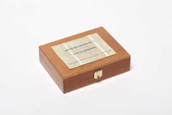 A wooden box with a white label on the lid with blue print "Boite Optimste No. 2 Vive le...