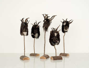 Artist created versions of Krampus Masks, each displayed on a single branch secured into a log.