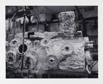 A black and white image of a foil covered piece of scientific equipment.  The artist describes...