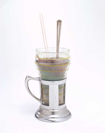 A soda glass and holder filled with blue wax, hair, and meat-like chunks.  A spoon and straw are...