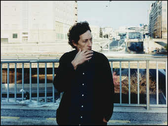 A man wearing a blue coat, standing in front of an urban landscape, with his hand touching the...