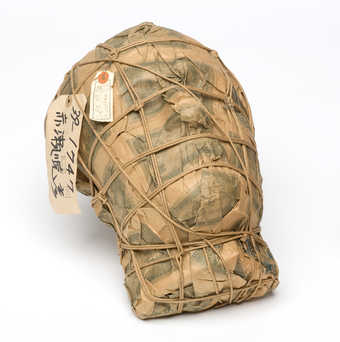 A plaster mask covered with imitation one-thousand-Yen notes, wrapped with a string netting. ...